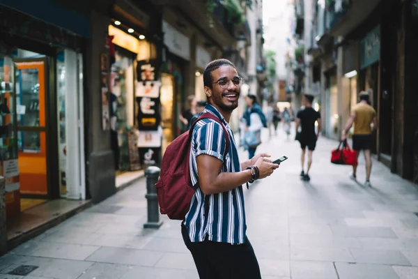 幸せなアフリカ系アメリカ人男性の観光客は笑って 休日の休暇にスマートフォンで街の通りを歩いて 正の暗い肌のヒップスター男は旅の途中で携帯電話でカメラを散歩見て — ストック写真