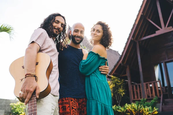 Neşeli çoklu etnik arkadaşlardan oluşan düşük açılı bir grup güler yüzlü, gitarla takılan, kucaklaşan ve kameraya bakan.
