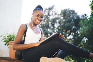 Gözlüklü neşeli kadın blog yazarının portresi kameraya gülümseyen ve kulaklıklı, kulaklıklı, kulaklık takan mutlu Afrikalı Amerikalı bir kadının müzik listesini dinlerken dokunmatik ped uygulaması kullanıyor.