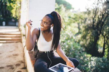 Elektronik kulaklıklı koyu tenli bayan öğrenci müzik podcast 'i ile boş zaman geçiriyor dijital tablette medya uygulaması kullanıyor, dokunmatik ped kullanan ve açık havada gülümseyen neşeli hippi kız.