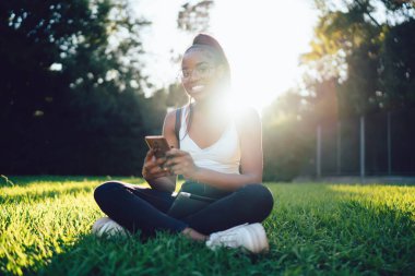 Güneşli parkta boş zamanlarında cep telefonu uygulaması kullanan, optik gözlüklü Afro-Amerikan yeniyetme kızın arka ışıklandırılmış portresi, akıllı telefonuyla kameraya gülümseyen neşeli kadın blogcu