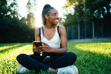 Neşeli Afrikalı Amerikalı hippi kız görme düzeltmesi için gözlüklü güneşli parkta boş zamanlarında mobil uygulama kullanıyor, akıllı telefonlu neşeli kadın blogcu başka tarafa bakıyor ve gülümsüyor.