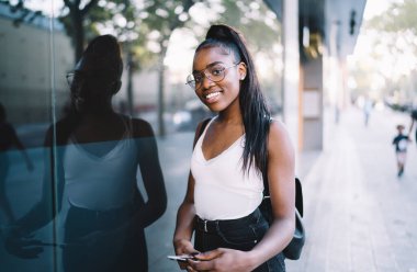 Optik gözlüklü mutlu koyu tenli kadının yarım boy portresi kameraya gülümseyen gözler sağlamak için şehir ortamında dokunarak, binlerce yıllık Afrikalı Amerikalı hippi kız pozu vererek.