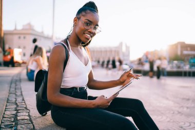 Modayı takip eden gözlüklü mutlu hippi kızın portresi. Seyahat sırasında dokunmatik ped cihazı kullanıyor. Cihaz teknolojisi olan mutlu esmer tenli turist şehirde dinlenmek için kameraya gülümsüyor.