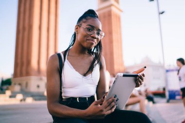 Genç hippi kızın portresi, elinde modern bir dokunmatik ped tutarken ve şehirde dinlenirken kameraya bakarken, açık havada dijital tabletle gezen koyu tenli seyahat blogcusu.