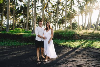 Yaz kıyafetleri içinde birbirini okşayan uzun bir çift kumlu sahilde avuç içleriyle vakit geçirip kameraya bakıyor.