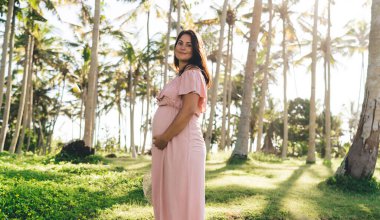 Yan görünüşlü, hamile, pembe elbiseli, çıplak omuzlu, karnını okşayan ve güneşli bir günün keyfini çıkarırken gülümseyen, gülümseyen bir kadın.