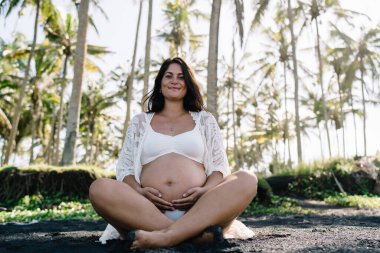 Düşük açılı mutlu, hamile, bikinili kadın gezgin, gülümseyen ve Lotus pozunda dinlenirken kameraya bakan kumlu tropikal sahilde meditasyon yapan.