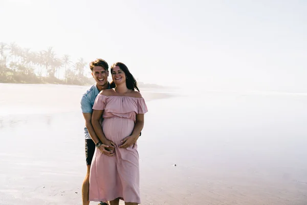 Felice Giovane Uomo Sorridente Abbracciando Allegra Moglie Incinta Mentre Piedi — Foto Stock