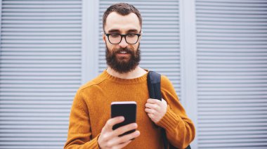 Gözlüklü ve sırt çantalı sakin erkek geniş açılı. Duvarın yanında duruyor. Akıllı telefondan video çağrısı alıyor.