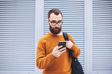 Sırt çantalı sakallı bir erkek sokakta duruyor ve sosyal ağların boş zamanlarını kontrol etmek için akıllı telefon kullanıyor.