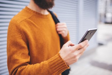 Sırt çantalı yüzsüz bir erkek duvarın yanında duruyor dinlenirken cep telefonuna bakıyor.