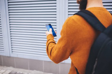 Koyu sarı kazaklı ve siyah sırt çantalı tanınmayan sakallı bir adam binanın yanında camları silindirli bir cep telefonu çeviriyor.