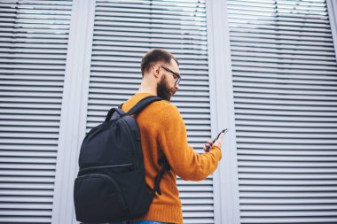 Düşük açılı, ince sakallı, koyu sarı gözlüklü hippi ve siyah sırt çantalı gezici cep telefonu pencerelerinde perdeleri olan binanın yanında duruyor.