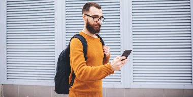Moda giysileri içindeki genç hipster 'ın yan görüntüsü ve siyah sırt çantasıyla sokakta cep telefonuyla mesaj yolluyor.