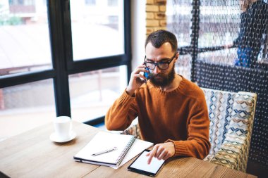 Akıllı telefonla konuşurken tablet ekranına dokunan ve gündüzleri kafede telgraf çekerken not alan sakallı adam.