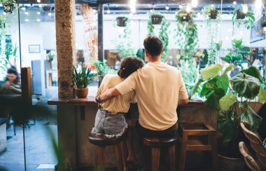 Tarz sahibi giysiler içinde tanınmayan genç bir adam ve kadının, yeşil bitkilerle süslenmiş rahat bir kafede birlikte vakit geçirirken sarılışının arka planında.