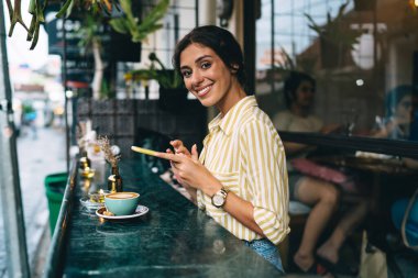 Kafe terasında köpüklü kapuçino ile otururken şık giyinmiş, cep telefonu kullanan genç bir kadının yan görüntüsü.