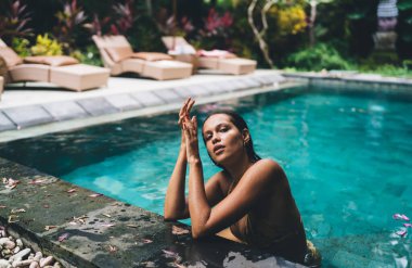 Mayo giymiş yüksek açılı bir kadın yaz tatilinde yüzme havuzunda dinlenirken kameraya bakıyor ve dinleniyor.