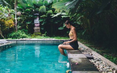 Yüzme havuzunda otururken bilgi edinmek için dizüstü bilgisayar kullanan mayo ve tişörtlü genç kadının yan görüntüsü