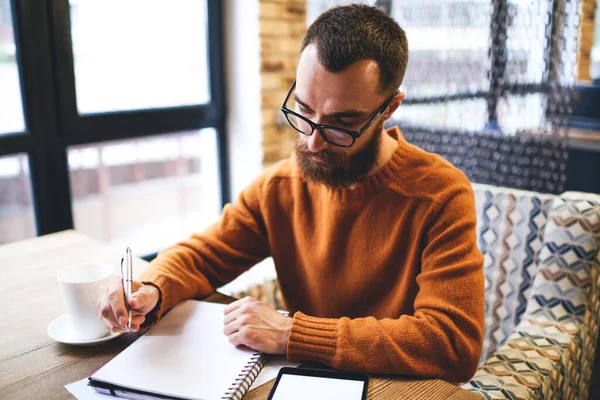 从上图可以看出 留胡子的男作家穿着休闲装 戴着眼镜 一边在笔记本上写文章 一边在舒适的咖啡馆里喝杯茶 一边吃平板电脑 — 图库照片