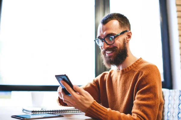 Vousatý Muž Zubatým Úsměvem Sedí Stolu Dívá Obrazovku Mobilního Telefonu — Stock fotografie