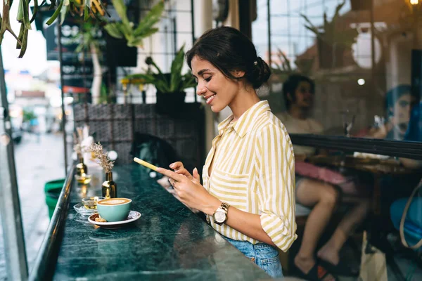 カプチーノを飲みながらコーヒーブレイクをしたり スタイリッシュなカフェテラスでスマートフォンを使用する流行の衣装で暗い髪を持つ肯定的な若い民族女性の側面図 — ストック写真