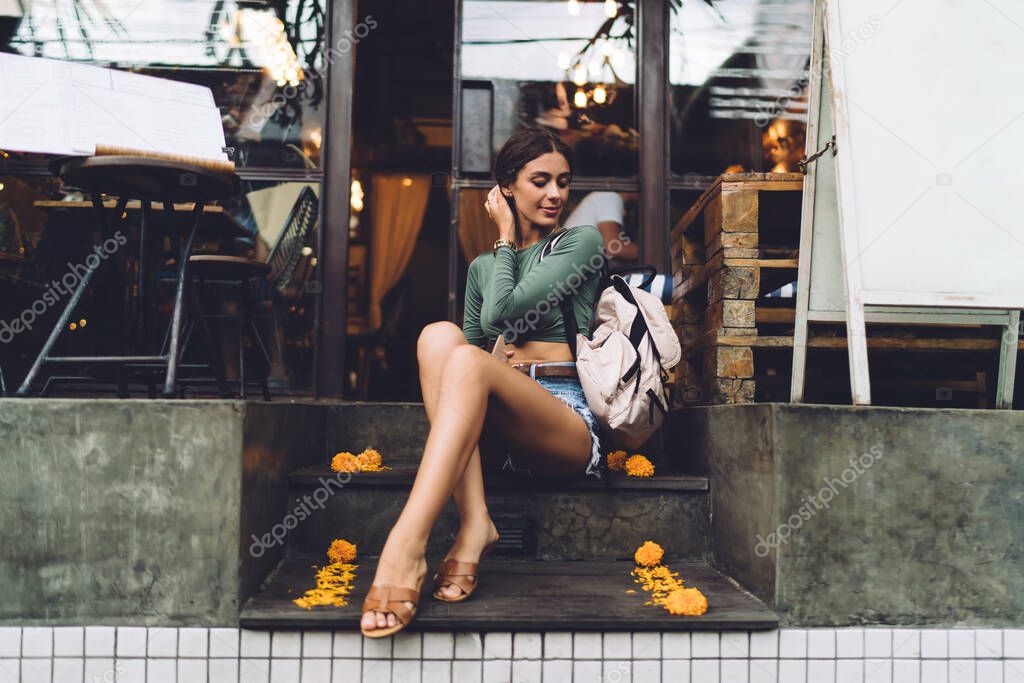 Full body of slim female model in casual outfit resting on stone stairs near glass doorway of building and touching hair while looking down