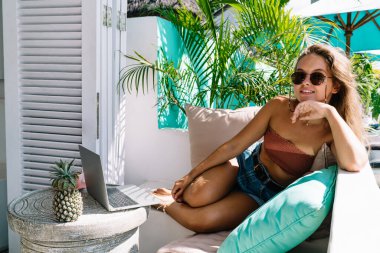 Rahat koltukta uzanan ve Bali tatil beldesinde yaz tatilinin tadını çıkaran, gündelik kıyafet ve güneş gözlüklü tam vücut içerikli bir kadın.