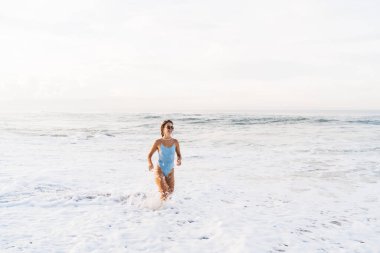 Şık mavi mayo giymiş mutlu, ince kadın köpüklü denizde kıyıda duruyor ve güzel havayı seyrediyor.