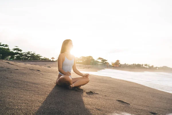 Çıplak ayaklı, mayolu bir kadın. Gün batımında meditasyon yaparken sakin deniz suyunun yanındaki kumlu sahilde dinleniyor.