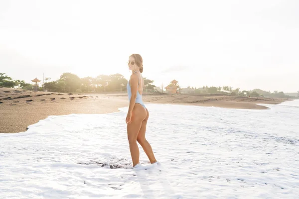 サイドビュー魅力的な女性完璧なボディを身に着けているスタイリッシュな水着とサングラス砂浜に立って泡の海の波に洗われ カメラを見て — ストック写真