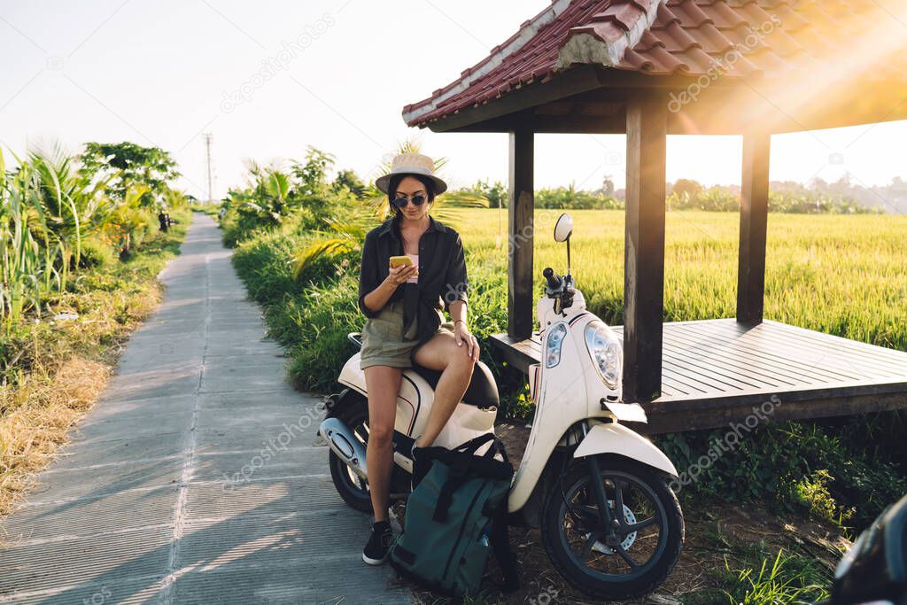 Millennial female user checking location information during online GPS tracking via modern cellphone gadget, travel girl resting on rent moped connecting to roaming internet for browsing website