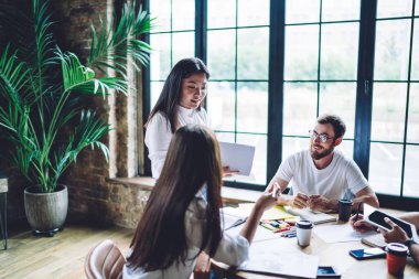 Beyaz tişörtlü neşeli çok ırklı takım üyeleri masada oturmuş, açık modern işyerinde birlikte çalışırken yeni projeden bahsediyorlar.