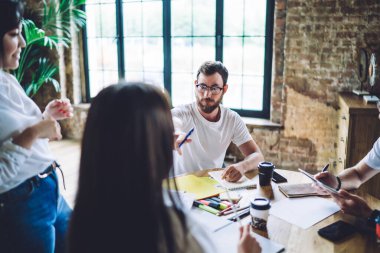 Yüksek açılı, genç, sakallı, isimsiz bir kadını işaret eden, yaratıcı stüdyodaki çok ırklı iş arkadaşlarıyla masaya oturmuş, kahve ve not defterleriyle beyin fırtınası yapan,