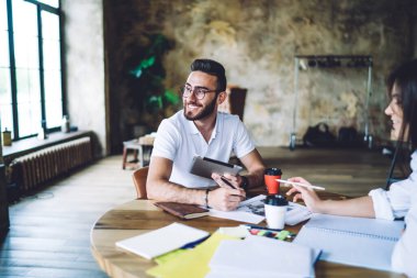 Çatı katında çalışırken tablet kullanan ve yuvarlak masa başında not alan neşeli, yaratıcı genç adam ve kadın.