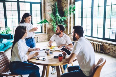 Beyaz gömlekli, kot pantolonlu, zeki Asyalı bir bayanın, yaratıcı ofiste toplantı sırasında sofrada oturan etnik çeşitliliğe sahip iş arkadaşlarına, ellerinde kahve ve aletlerle görüşlerini açıklamasıyla yüksek açı.