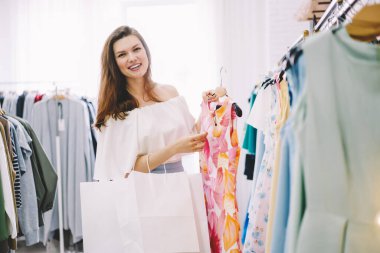 Beyaz elbiseli neşeli esmer, beyaz alışveriş torbalarıyla kameraya bakıyor ve moda mağazasından alışveriş için elbise seçerken gülümsüyor.