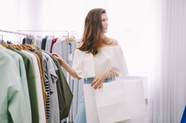 Beyaz alışveriş torbaları tutan ve gündüzleri şık bir butikte kıyafet seçen alımlı bayanın yan görüntüsü.