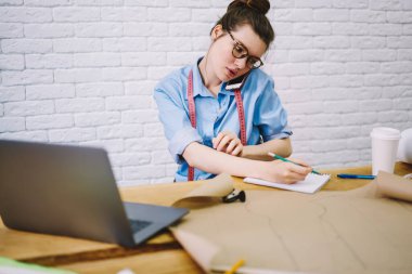 Günlük kıyafetli genç bir kadın terzi ve boynundaki bantla akıllı telefonla konuşuyor ve atölyedeki yeni düzenin detaylarını not alıyor.