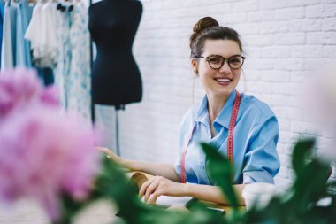 Moda stüdyosunda çalışırken boynunda gözlük ve mezurayla kameraya bakan mutlu bir terzi.