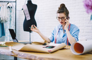 Gayri resmi giyinen ve boynundaki bantla genç bayan moda tasarımcısı yeni projenin cep telefonu detaylarını tartışıyor kumaş örnekleri ve çizimlerle masada oturuyor.