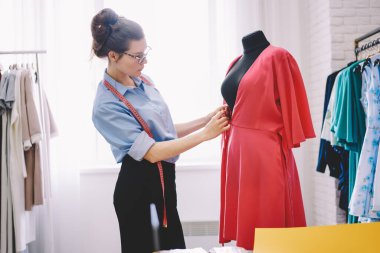 Düşünceli kadın terzinin, ölçüm bandıyla mankenin yanında durup ölçü alırken modern atölyede yeni moda elbiseler dikerken görüntüsü.