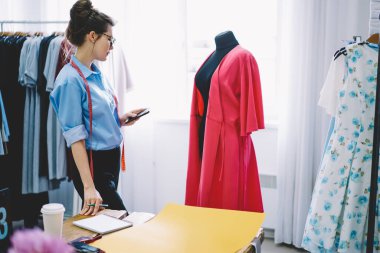Modern hafif iş yerinde çalışırken akıllı telefon kullanan parlak elbiseli mankenin yanında duran düşünceli kadın terzinin yan görüntüsü