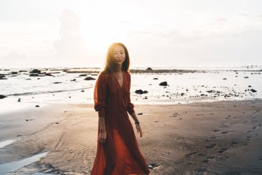 Bali 'de kumlu sahilde gezinen, terracotta yaz elbisesi giymiş genç, narin bir kadın.