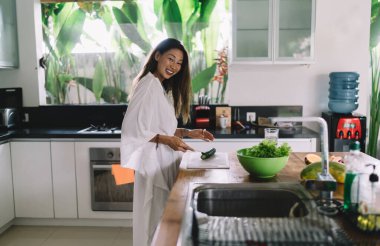 Beyaz cüppeli, şık Asyalı bir bayanın yemek hazırlığı sırasında gülümseyip kameraya bakarken salatalık keserken görüntüsü.