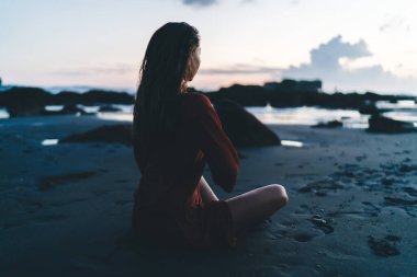 Kırmızı elbiseli Asyalı bir kadın deniz kenarındaki kayalık sahilde oturuyor ve gözlerini kaçırarak güneşin doğuşunu seyrediyor.
