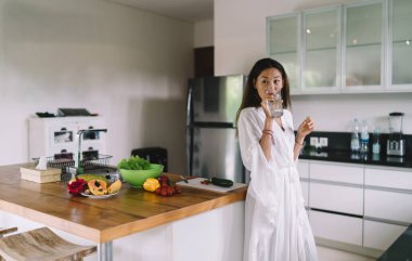 Beyaz cüppeli, uzun siyah saçlı, genç, sıska bir Asyalı kadın. Bir yandan mutfak masasının yanında çeşitli meyve ve sebzelerle dikilirken bir yandan da bir bardak su içiyor.