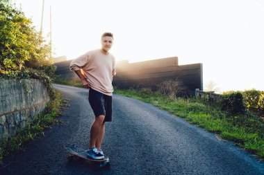 Arka taraftaki genç sporcu, bir ayağı kaykayın üstünde kırsal alanda kaldırım kenarında duruyor ve gün batımında kameraya bakıyor.