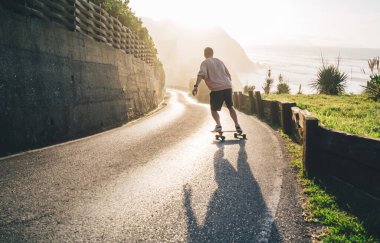 Yazın asfalt sokakta, parlak güneşin altında şortlu ve svetşörtlü, kendinden emin bir adamın uzun tahta sürüşünün arka görüntüsü.